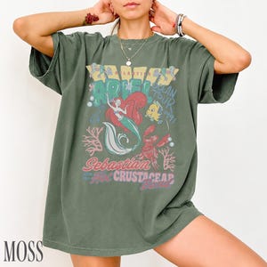 Puede incluir: Una camiseta de manga corta color verde musgo con un estampado gr&aacute;fico de Ariel, Flounder y Sebasti&aacute;n. El texto dice Ariel Ocean Tour 1991, Sebastian and the Hot Crustacean Band. El dise&ntilde;o incluye corales y burbujas en tonos rojos, azules y blancos.
