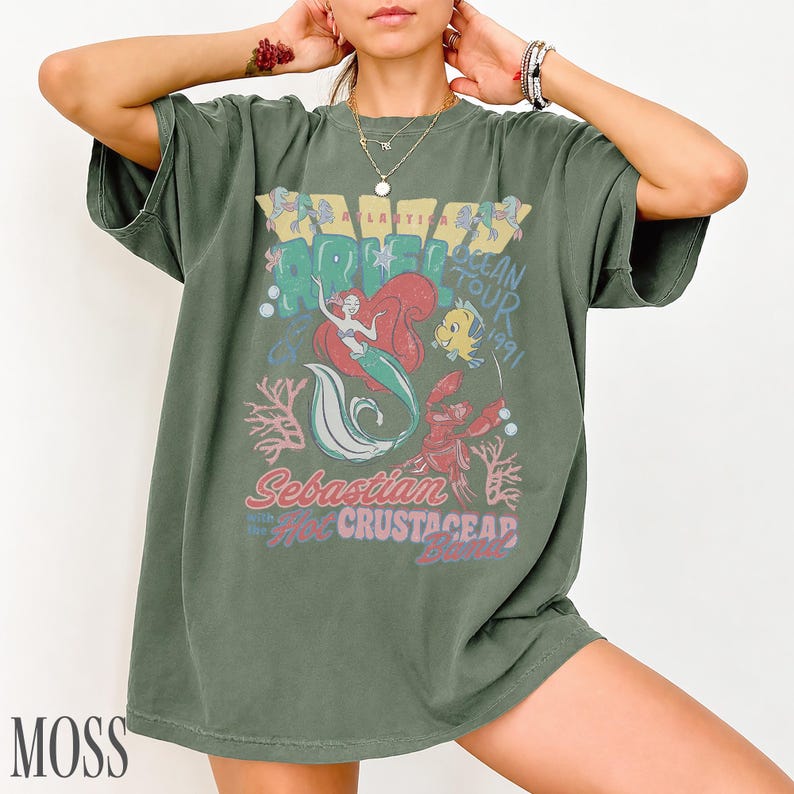 Puede incluir: Una camiseta de manga corta color verde musgo con un estampado gr&aacute;fico de Ariel, Flounder y Sebasti&aacute;n. El texto dice Ariel Ocean Tour 1991, Sebastian and the Hot Crustacean Band. El dise&ntilde;o incluye corales y burbujas en tonos rojos, azules y blancos.
