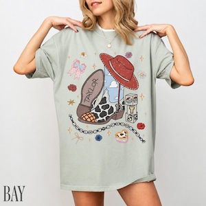 May include: A sage green t-shirt featuring a graphic print of a cowboy boot with a cow print pattern, a red hat, a beaded necklace, and blocks spelling out TS. The boot has the name Taylor written on the sole and the number 13 on the side.