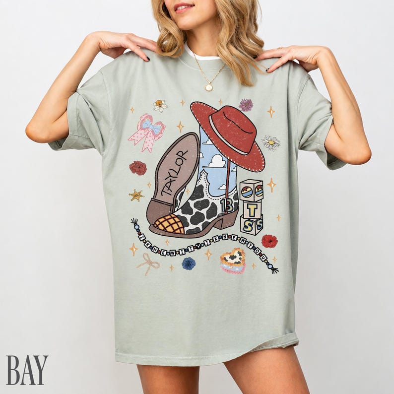 May include: A sage green t-shirt featuring a graphic print of a cowboy boot with a cow print pattern, a red hat, a beaded necklace, and blocks spelling out TS. The boot has the name Taylor written on the sole and the number 13 on the side.