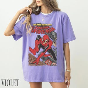 May include: Violet t-shirt featuring a comic book graphic of Spider-Man. The design includes the text "BRAND NEW DAY" and "AMAZING SPIDER-MAN". The superhero is depicted in a dynamic pose against a cityscape backdrop. The word "VIOLET" is printed at the bottom.