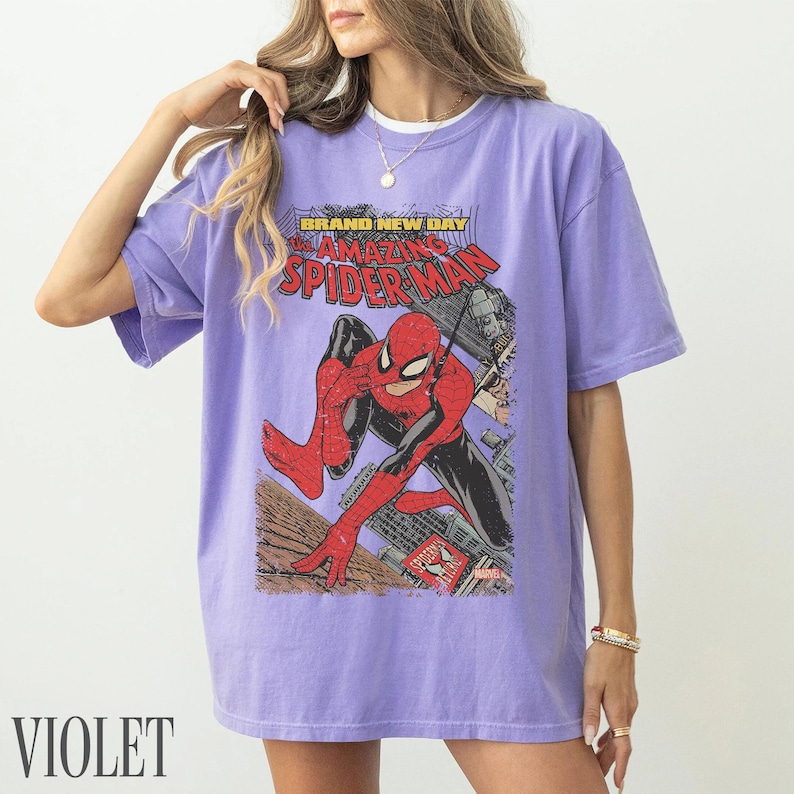 May include: Violet t-shirt featuring a comic book graphic of Spider-Man. The design includes the text "BRAND NEW DAY" and "AMAZING SPIDER-MAN". The superhero is depicted in a dynamic pose against a cityscape backdrop. The word "VIOLET" is printed at the bottom.