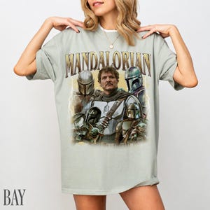 May include: A pale green t-shirt with a retro graphic of characters from The Mandalorian. The design includes the word "MANDALORIAN" in a gold, distressed font, and images of the characters. The shirt has a loose fit.