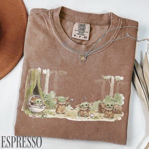 Pu&ograve; includere: T-shirt color espresso con una scena della foresta con personaggi Grogu. La maglietta ha un girocollo e maniche corte. Una collana d'argento con un ciondolo &egrave; drappeggiata sulla maglietta. La parola "ESPRESSO" &egrave; stampata in basso.