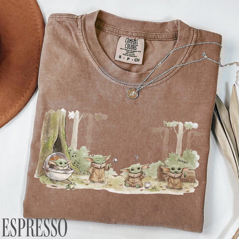 Pu&ograve; includere: T-shirt color espresso con una scena della foresta con personaggi Grogu. La maglietta ha un girocollo e maniche corte. Una collana d'argento con un ciondolo &egrave; drappeggiata sulla maglietta. La parola "ESPRESSO" &egrave; stampata in basso.