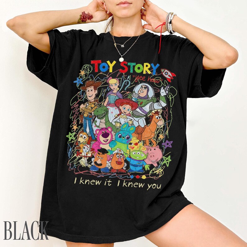 May include: A black short-sleeved t-shirt featuring a colourful graphic print of Toy Story characters including Woody, Buzz Lightyear, and Jessie. The design includes scribbled star accents, the text "TOY STORY", "yee haw", and "I knew it I knew you".
