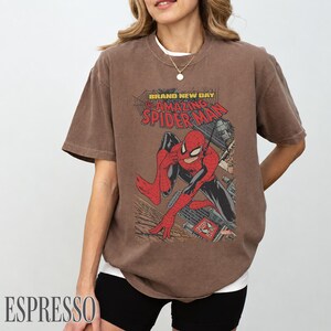 May include: Espresso-colored t-shirt featuring a comic book graphic of Spider-Man. The design includes the text "BRAND NEW DAY" and "THE AMAZING SPIDER-MAN". The superhero is depicted in a dynamic pose, with a cityscape backdrop.