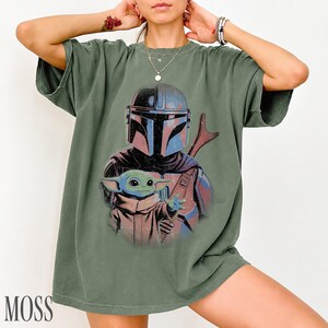 Puede incluir: Una camiseta de manga corta color verde oliva con un estampado gr&aacute;fico de un personaje con casco y armadura oscura detr&aacute;s de una peque&ntilde;a criatura verde de orejas y ojos grandes. El dise&ntilde;o tiene una est&eacute;tica desgastada y vintage.