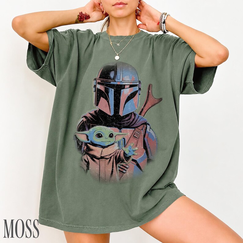 Puede incluir: Una camiseta de manga corta color verde oliva con un estampado gr&aacute;fico de un personaje con casco y armadura oscura detr&aacute;s de una peque&ntilde;a criatura verde de orejas y ojos grandes. El dise&ntilde;o tiene una est&eacute;tica desgastada y vintage.