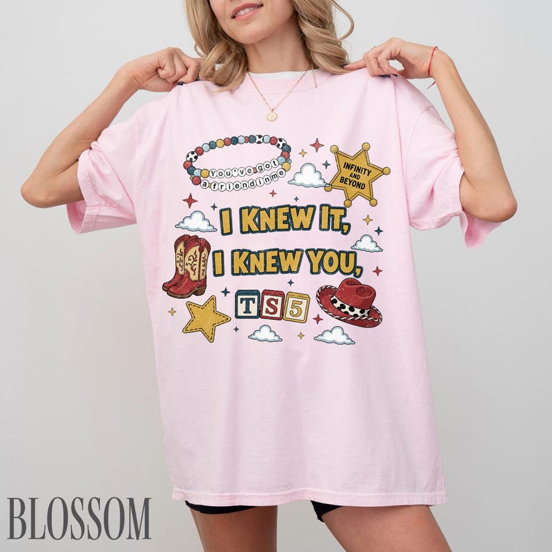 May include: A light pink t-shirt featuring a graphic print with a beaded bracelet, a gold sheriff star, cowboy boots, a red cowboy hat, and alphabet blocks spelling TS5. The text reads I KNEW IT, I KNEW YOU, INFINITY AND BEYOND.