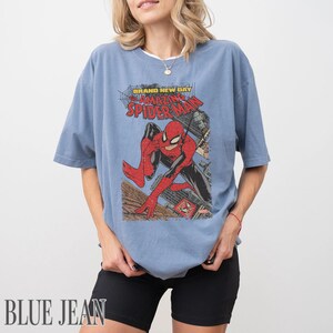 May include: Blue jean-colored t-shirt featuring a vintage comic book graphic of Spider-Man. The graphic includes the text "BRAND NEW DAY" and "AMAZING SPIDER-MAN". The shirt has short sleeves and a relaxed fit.