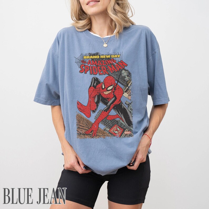 May include: Blue jean-colored t-shirt featuring a vintage comic book graphic of Spider-Man. The graphic includes the text "BRAND NEW DAY" and "AMAZING SPIDER-MAN". The shirt has short sleeves and a relaxed fit.