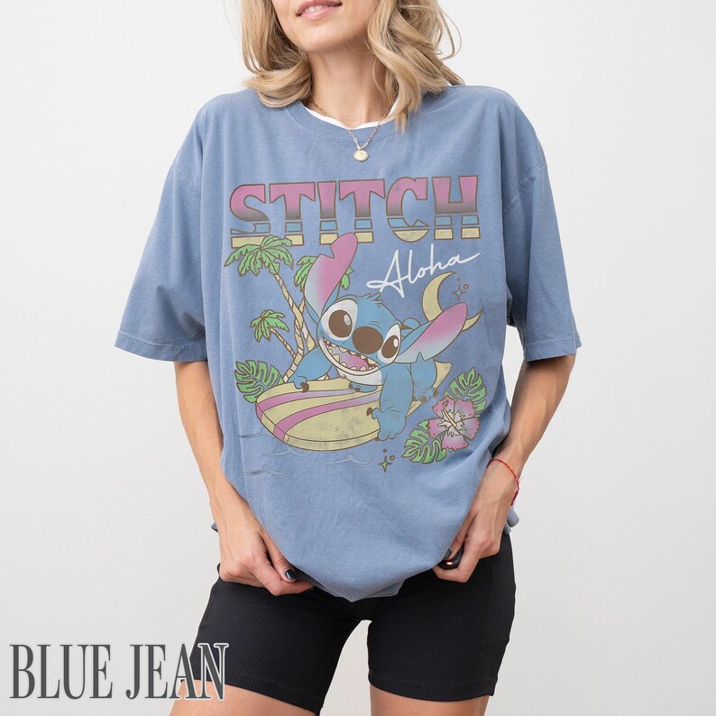 May include: A blue t-shirt featuring a graphic of Stitch surfing with the text "STITCH Aloha". The design includes palm trees and a pink flower. The shirt is a relaxed fit.