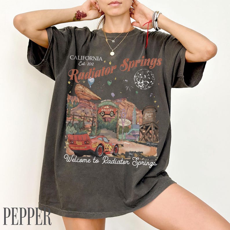 May include: A charcoal grey short-sleeve t-shirt featuring a graphic print of Radiator Springs with a red race car, desert rock formations, a water tower, and the text California Est. 2012 Radiator Springs Welcome to Radiator Springs.