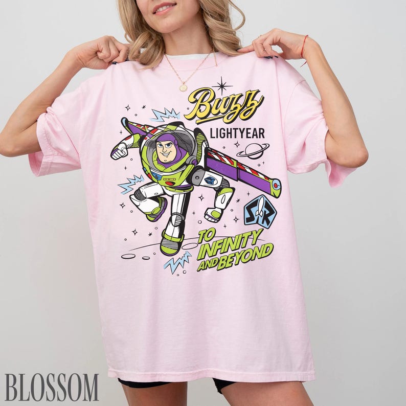Pode incluir: Camiseta rosa claro com um desenho do Buzz Lightyear, com o texto "Buzz Lightyear" e "To Infinity and Beyond". O desenho mostra Buzz Lightyear em seu traje espacial. A camiseta tem mangas curtas.