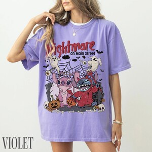 May include: A light purple short-sleeve t-shirt featuring a graphic print with the text Nightmare on Main Street. The design includes two cartoon characters, one pink and one blue, surrounded by ghosts, spiderwebs, pumpkins, and skulls.