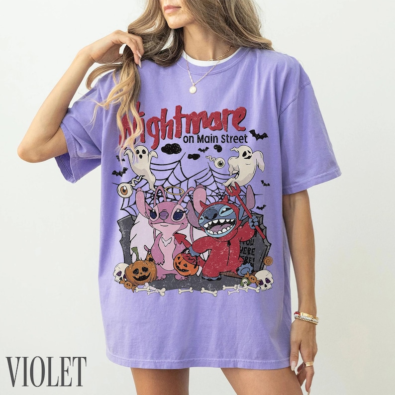 May include: A light purple short-sleeve t-shirt featuring a graphic print with the text Nightmare on Main Street. The design includes two cartoon characters, one pink and one blue, surrounded by ghosts, spiderwebs, pumpkins, and skulls.