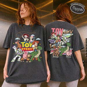 K&ouml;nnte beinhalten: Dunkelgraues T-Shirt mit einem farbenfrohen Toy Story-Motiv auf der Vorder- und R&uuml;ckseite. Das Frontdesign zeigt Woody, Buzz Lightyear und andere Charaktere. Das R&uuml;ckendesign zeigt "Toy Story" mit dem Text "Jessie Since 1995".