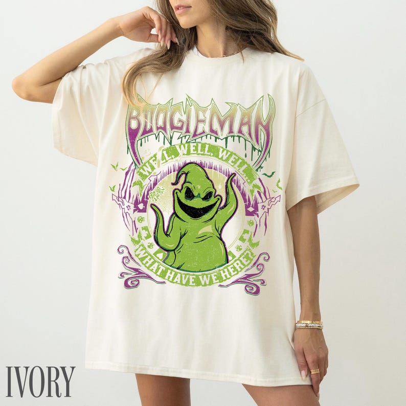 May include: Ivory-coloured t-shirt with a graphic of Oogie Boogie from The Nightmare Before Christmas. The design includes the text "BOOGIEMAN" and "WE'LL WELL WELL! WHAT HAVE WE HERE?" in green and purple.