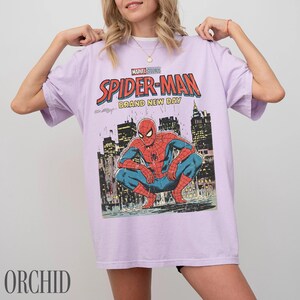 May include: A light purple t-shirt featuring a Spider-Man graphic. The graphic includes the words "Marvel Studios Spider-Man Brand New Day" and an illustration of Spider-Man crouching in front of a city skyline.