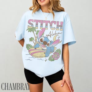 May include: Light blue t-shirt featuring a graphic of Stitch surfing with the text "STITCH Aloha" in pink and yellow. The design includes palm trees, a crescent moon, and a pink flower. The shirt is a casual wear item.