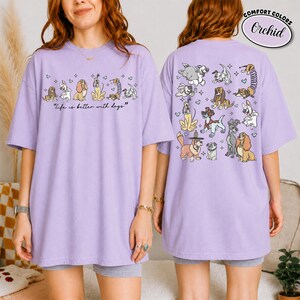Puede incluir: Una camiseta lila con un dise&ntilde;o de perros. La parte delantera muestra una l&iacute;nea de perros ilustrados con el texto "Life is better with dogs". La parte trasera de la camiseta tiene un gr&aacute;fico m&aacute;s grande de varias razas de perros, con la palabra "Orchid".