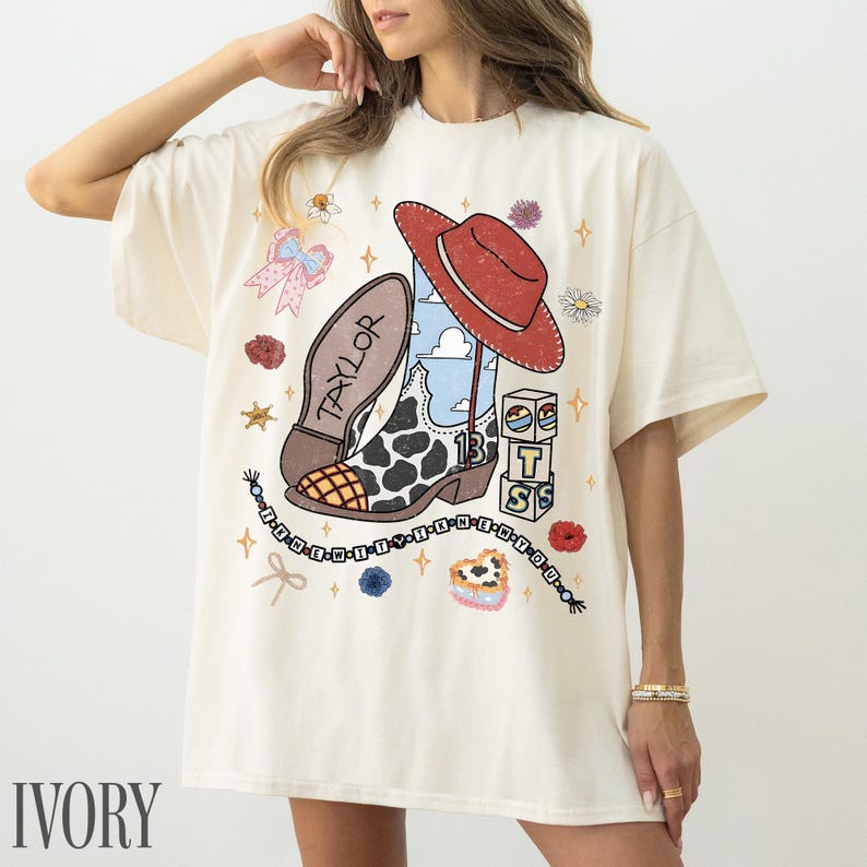 May include: An ivory short-sleeve t-shirt featuring a graphic print of a cowboy boot with a cow print pattern, a red hat, and the name Taylor. The design includes blocks with the letters T, S, and a sun icon, plus a beaded string with text.