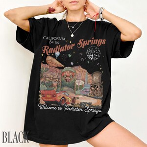 May include: A black t-shirt featuring a graphic print of a red race car in a desert landscape. The text reads California Est. 2012, Radiator Springs, and Welcome to Radiator Springs. The design includes a water tower, rock formations, and a moon.