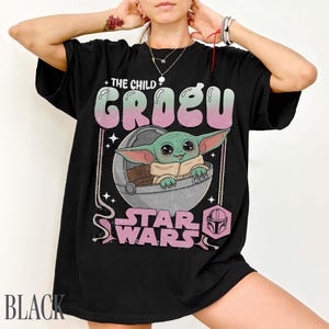 May include: A black t-shirt featuring a graphic print of Grogu from Star Wars sitting in a pod. The text reads THE CHILD GROGU and STAR WARS in a retro-style font. The design includes pink and green accents, small stars, and a helmet icon.