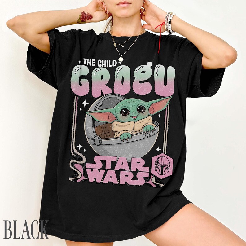 May include: A black t-shirt featuring a graphic print of Grogu from Star Wars sitting in a pod. The text reads THE CHILD GROGU and STAR WARS in a retro-style font. The design includes pink and green accents, small stars, and a helmet icon.