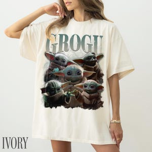 K&ouml;nnte beinhalten: Elfenbeinfarbenes T-Shirt mit einem Grogu-Motiv, mit dem Wort "GROGU" &uuml;ber dem Bild. Die Grafik zeigt mehrere Grogu-Charaktere in verschiedenen Posen. Das Shirt hat eine lockere Passform.