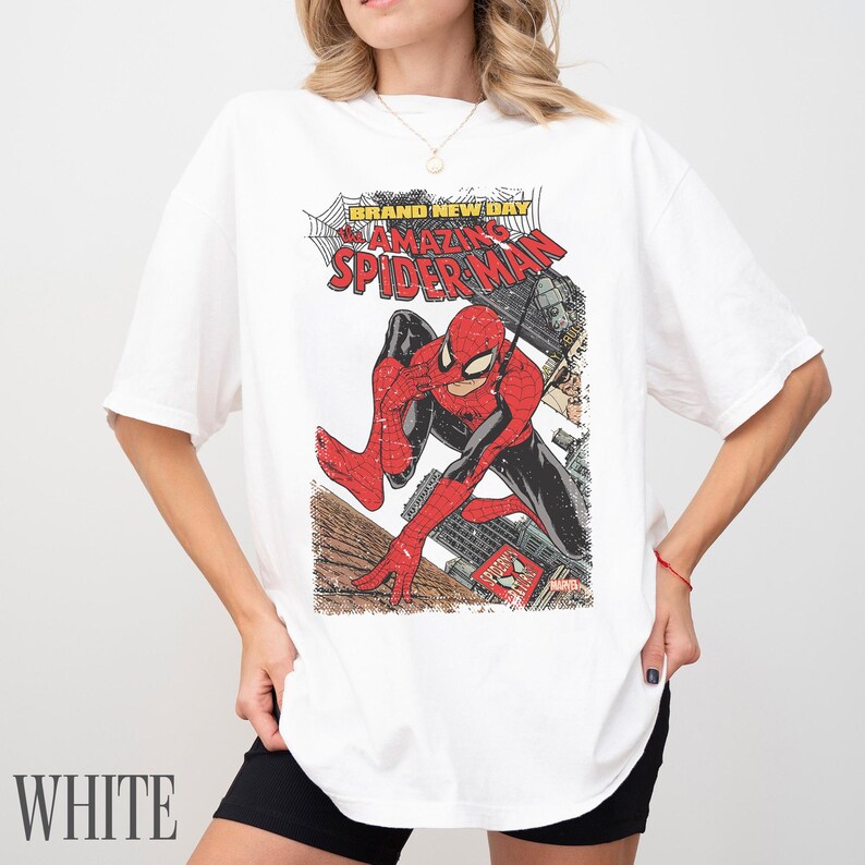 May include: White t-shirt featuring a comic book graphic of Spider-Man in a dynamic pose. The graphic includes the text "BRAND NEW DAY" and "AMAZING SPIDER-MAN". The shirt has short sleeves and a relaxed fit.