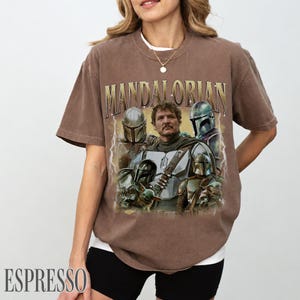 May include: Espresso-coloured t-shirt featuring a graphic of characters from the Mandalorian series. The design includes the text "MANDALORIAN" in a gold font. The shirt has a crew neck and short sleeves.