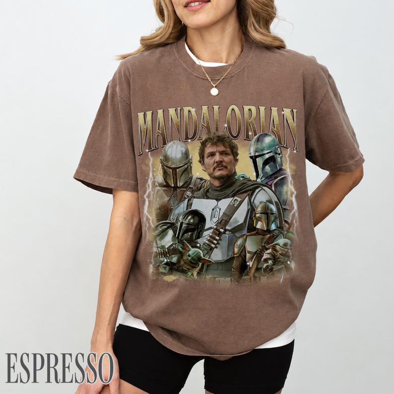 May include: Espresso-coloured t-shirt featuring a graphic of characters from the Mandalorian series. The design includes the text "MANDALORIAN" in a gold font. The shirt has a crew neck and short sleeves.