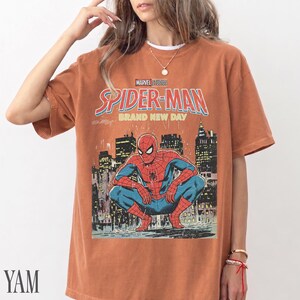 May include: A rust-colored t-shirt featuring a graphic of Spider-Man in a crouching pose against a cityscape backdrop. The shirt displays the text "MARVEL STUDIOS SPIDER-MAN BRAND NEW DAY" in bold lettering. The shirt has short sleeves.
