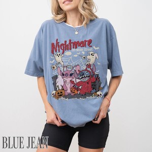 May include: A dusty blue short-sleeve t-shirt featuring a graphic print with the text Nightmare on Main Street. The design includes two cartoon characters, one pink and one blue, dressed in Halloween costumes with ghosts, pumpkins, and a spiderweb.