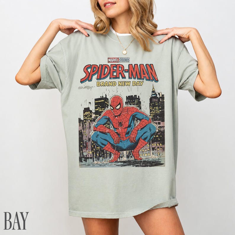 May include: A light green t-shirt featuring a Spider-Man comic book graphic. The graphic includes the words "Marvel Studios Spider-Man Brand New Day" and an illustration of Spider-Man in a city setting.