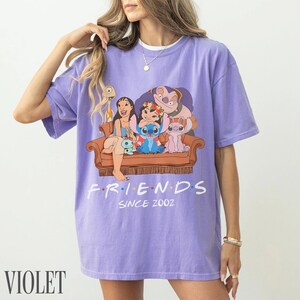May include: Lilac t-shirt with a cartoon image of Lilo & Stitch characters on a brown sofa. The word "FRIENDS" is above "SINCE 2002" in white. The word "VIOLET" is printed in white at the bottom. The t-shirt is a relaxed fit.