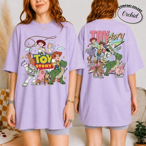 K&ouml;nnte beinhalten: Ein lila T-Shirt mit einer farbenfrohen Grafik von Charakteren aus dem Animationsfilm "Toy Story". Auf der Vorderseite des Shirts sind der Filmtitel und verschiedene Charaktere abgebildet, w&auml;hrend die R&uuml;ckseite weitere Charaktere und den Text "Since 1995" zeigt.
