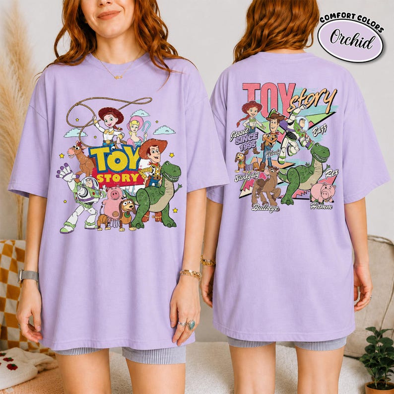 K&ouml;nnte beinhalten: Ein lila T-Shirt mit einer farbenfrohen Grafik von Charakteren aus dem Animationsfilm "Toy Story". Auf der Vorderseite des Shirts sind der Filmtitel und verschiedene Charaktere abgebildet, w&auml;hrend die R&uuml;ckseite weitere Charaktere und den Text "Since 1995" zeigt.