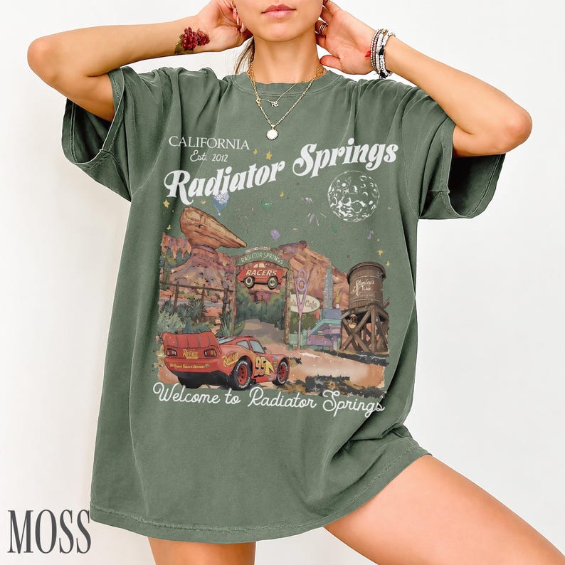 May include: An olive green short-sleeve t-shirt featuring a graphic print of Radiator Springs with a red race car, desert rock formations, a water tower, and the text California Est. 2012 Radiator Springs Welcome to Radiator Springs.