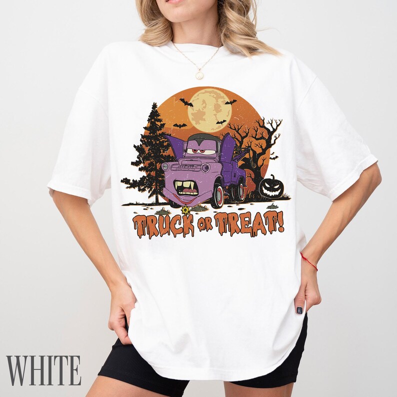 Może przedstawiać: Biała koszulka z grafiką na Halloween. Projekt przedstawia fioletową ciężar&oacute;wkę z cechami wampira, pełnię księżyca, nietoperze i napis "TRUCK OR TREAT!" w kolorze pomarańczowym i brązowym.