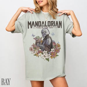 May include: A pale green t-shirt with the words "MANDALORIAN - THIS IS THE WAY" in dark grey. The design shows the Mandalorian and Grogu with flowers and butterflies.