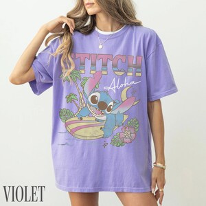 May include: Violet t-shirt featuring a graphic of Stitch surfing with the word "Stitch" above and "Aloha" below. The design includes palm trees, a surfboard, and tropical flowers. The shirt is a solid violet color.
