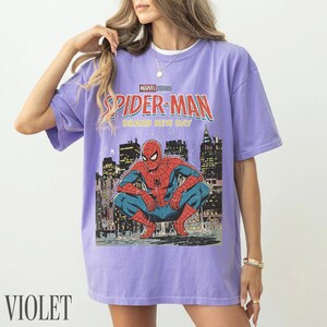 May include: Violet-colored t-shirt featuring a Spider-Man graphic. The design includes the text "Marvel Studios Spider-Man Brand New Day" above an illustration of Spider-Man crouching in front of a city skyline.