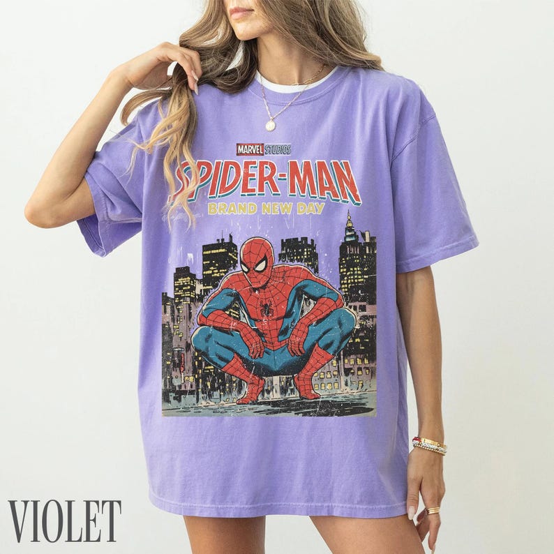 May include: Violet-colored t-shirt featuring a Spider-Man graphic. The design includes the text "Marvel Studios Spider-Man Brand New Day" above an illustration of Spider-Man crouching in front of a city skyline.
