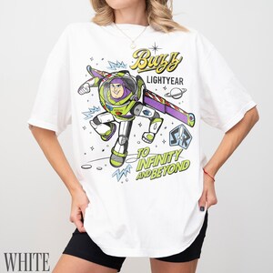 Pode incluir: Camiseta branca com um gr&aacute;fico colorido de Buzz Lightyear, o texto "Buzz Lightyear" e "To Infinity and Beyond". O design inclui um tema espacial com um planeta e estrelas. A camiseta tem um corte folgado.