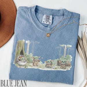 Pu&ograve; includere: T-shirt blu jeans con grafica di Grogu. La grafica presenta Grogu in un ambiente forestale. La maglietta ha girocollo e maniche corte. Una collana d'oro &egrave; drappeggiata sulla maglietta. La maglietta &egrave; etichettata "Comfort Colors".