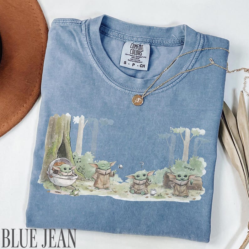 Pu&ograve; includere: T-shirt blu jeans con grafica di Grogu. La grafica presenta Grogu in un ambiente forestale. La maglietta ha girocollo e maniche corte. Una collana d'oro &egrave; drappeggiata sulla maglietta. La maglietta &egrave; etichettata "Comfort Colors".