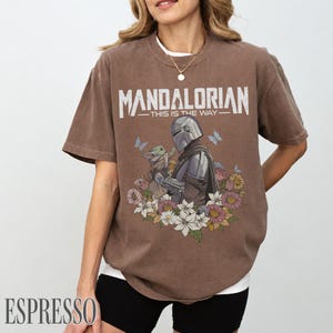 May include: Espresso-coloured t-shirt with the text "MANDALORIAN - THIS IS THE WAY" above an illustration of the Mandalorian and Grogu, surrounded by flowers and butterflies. The shirt has a relaxed fit and a white undershirt.